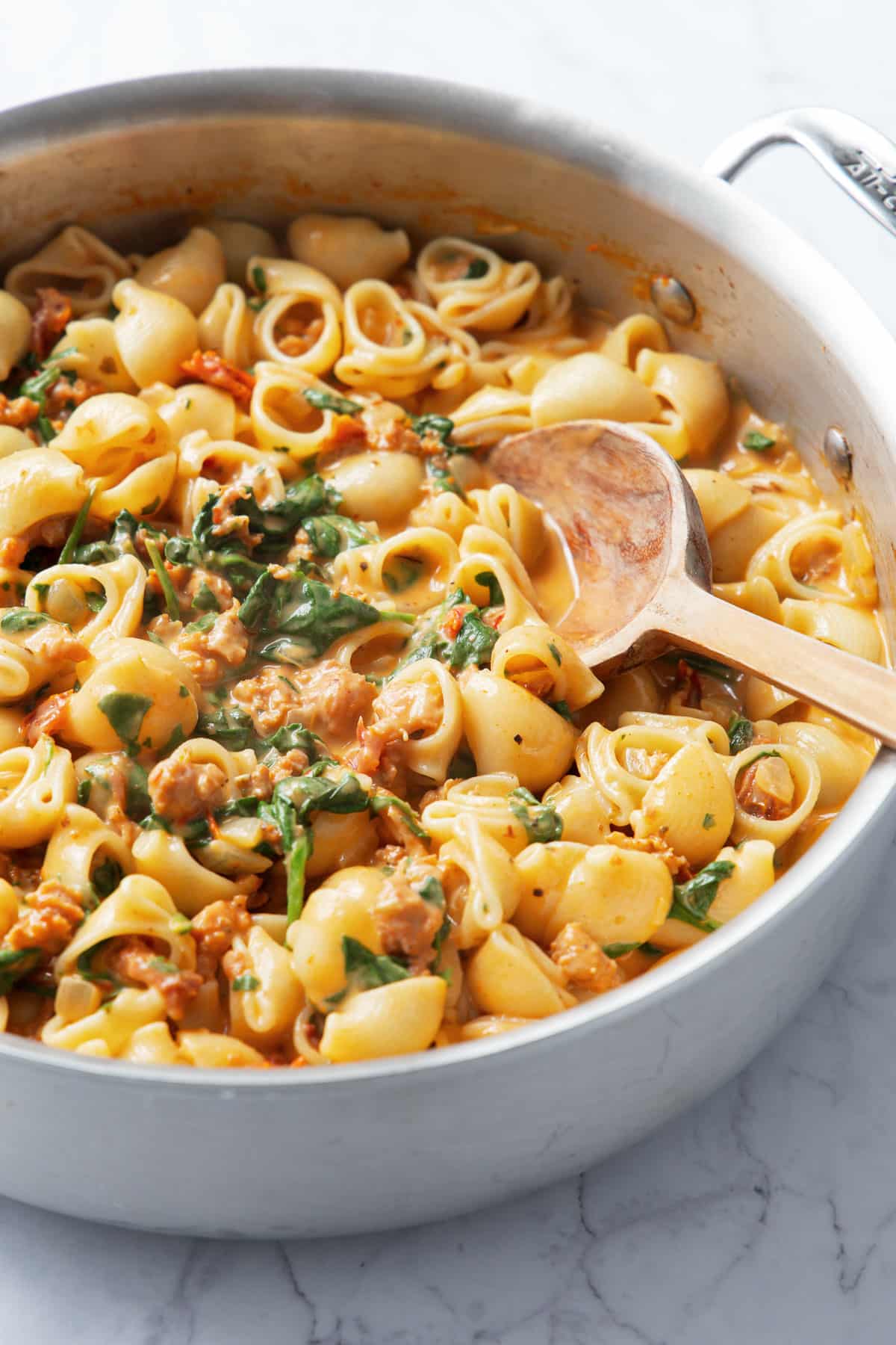 Closeup of stainless saucepan with Creamy Sundried Tomato and Italian Sausage Pasta and a wooden spoon.