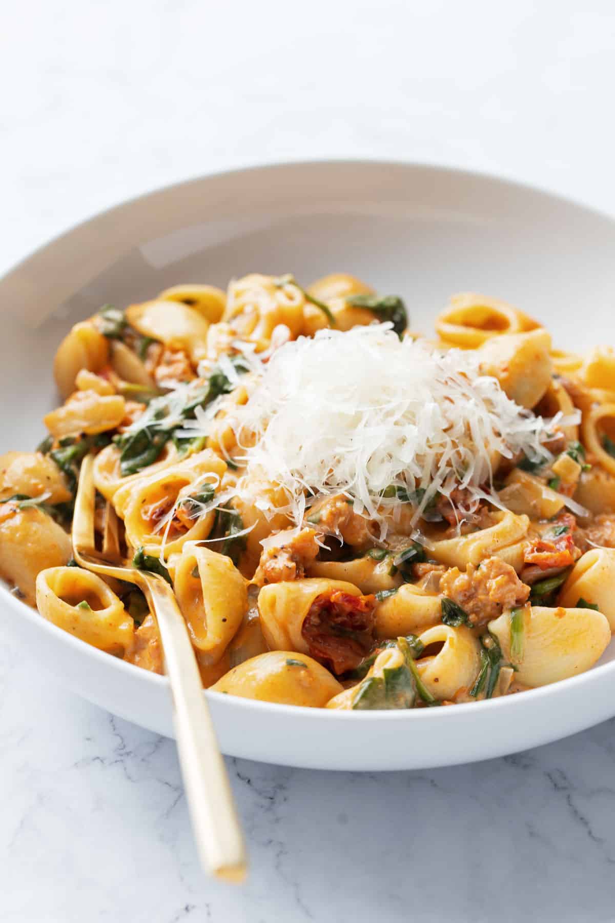 White bowl filled with Creamy Sundried Tomato and Italian Sausage Pasta and topped with finely shaved parmesan cheese with a gold fork.