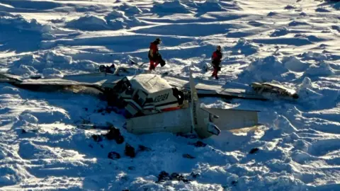 Reuters The wreckage of the plane, as seen from the air, surrounded by snow and debris as two people walk around it