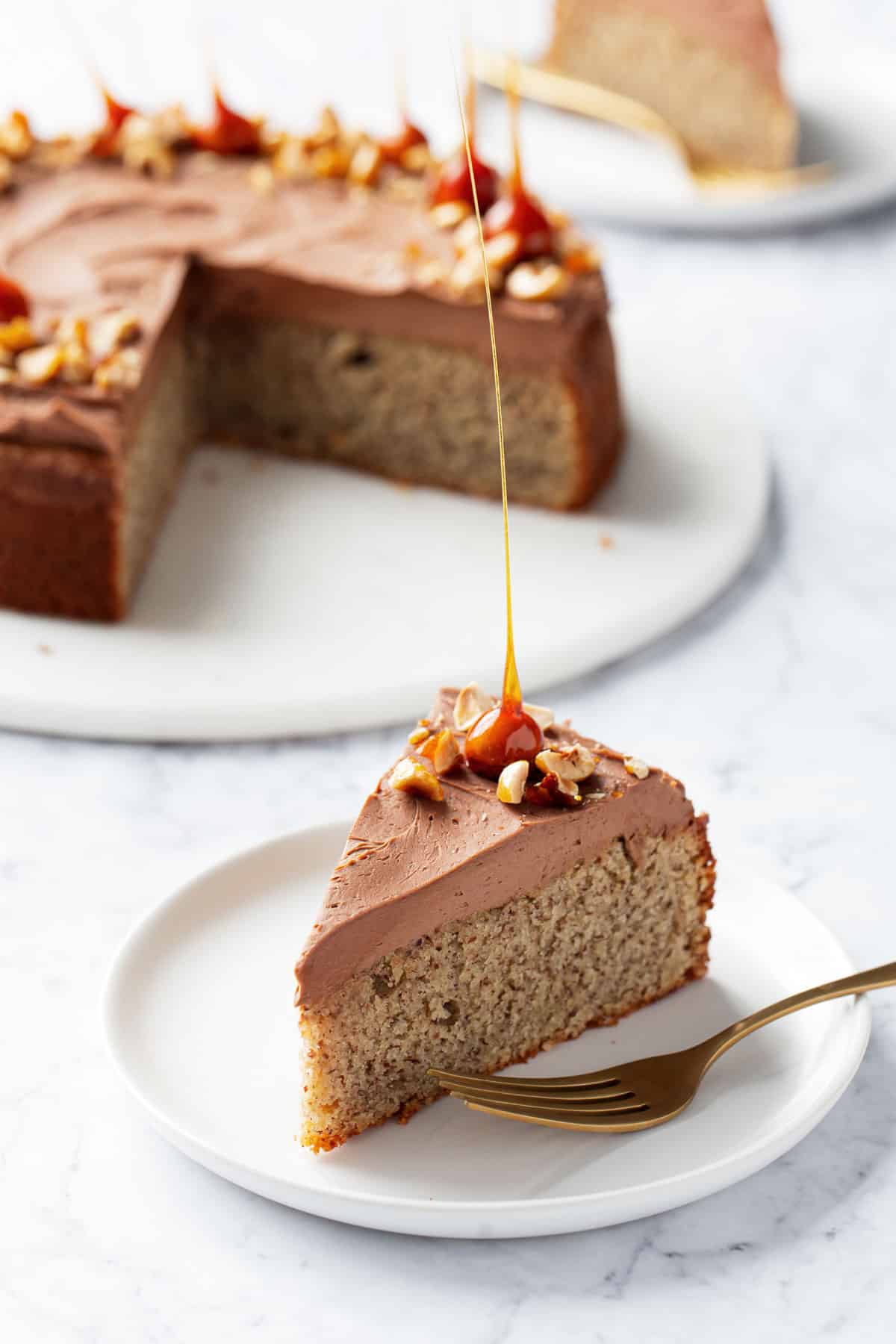 Slice of Flourless Hazelnut Cake with Nutella Whipped Ganache with gold fork on a white plate, with the rest of the cake on a serving plate in the background.