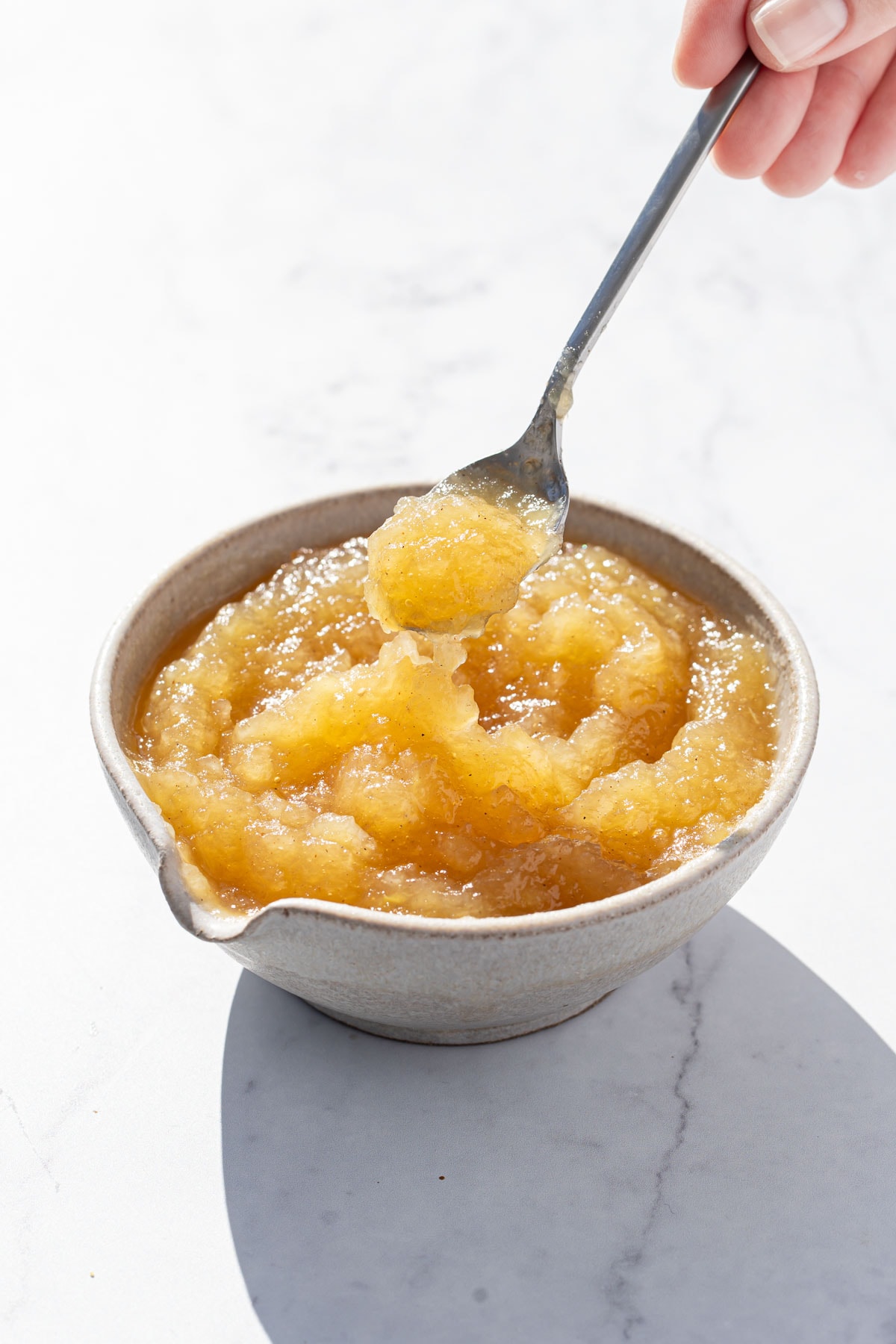 Spoonful of homemade applesauce lifting out of a bowl, in direct sunlight with harsh shadows on a white marble background.