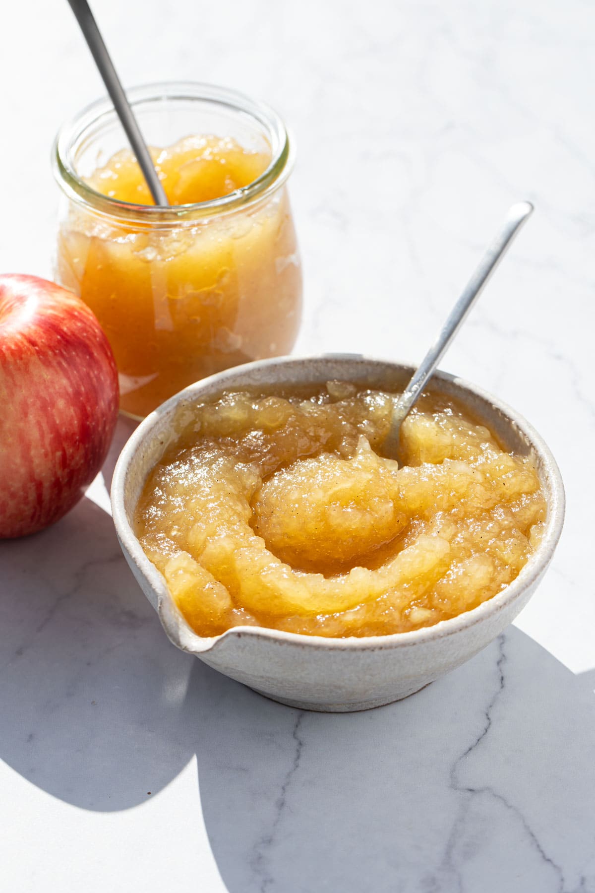 Bowl of homemade applesauce with a spoon, with red apple and a glass jar of applesauce off to the side.