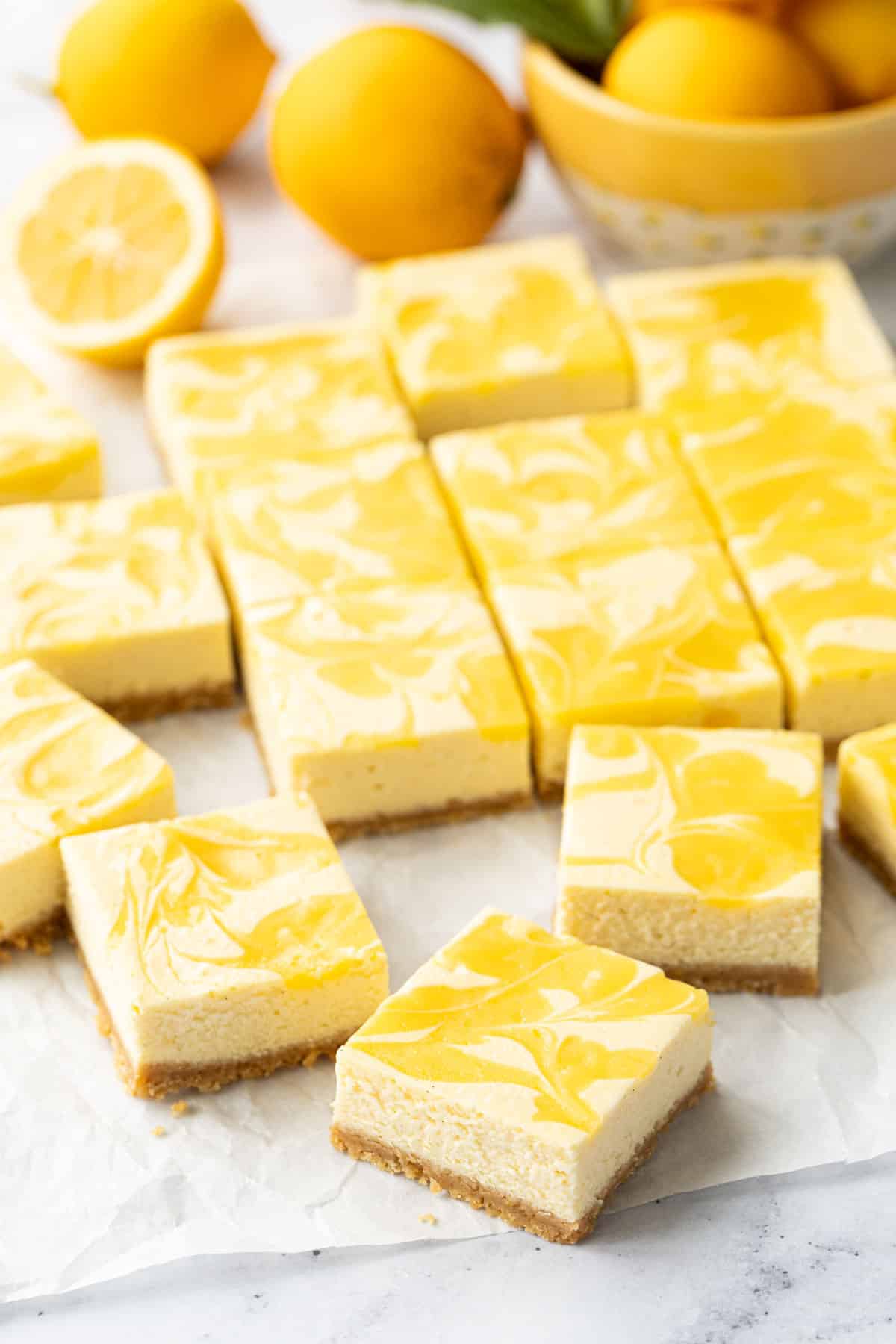 Cut squares of Meyer Lemon Cheesecake Bars arranged haphazardly on a piece of crinkled parchment paper, fresh lemons in the background.