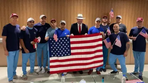 Embajada de los EE.UU., Venezuela Several men in jeans carrying a large American flag pose for photo