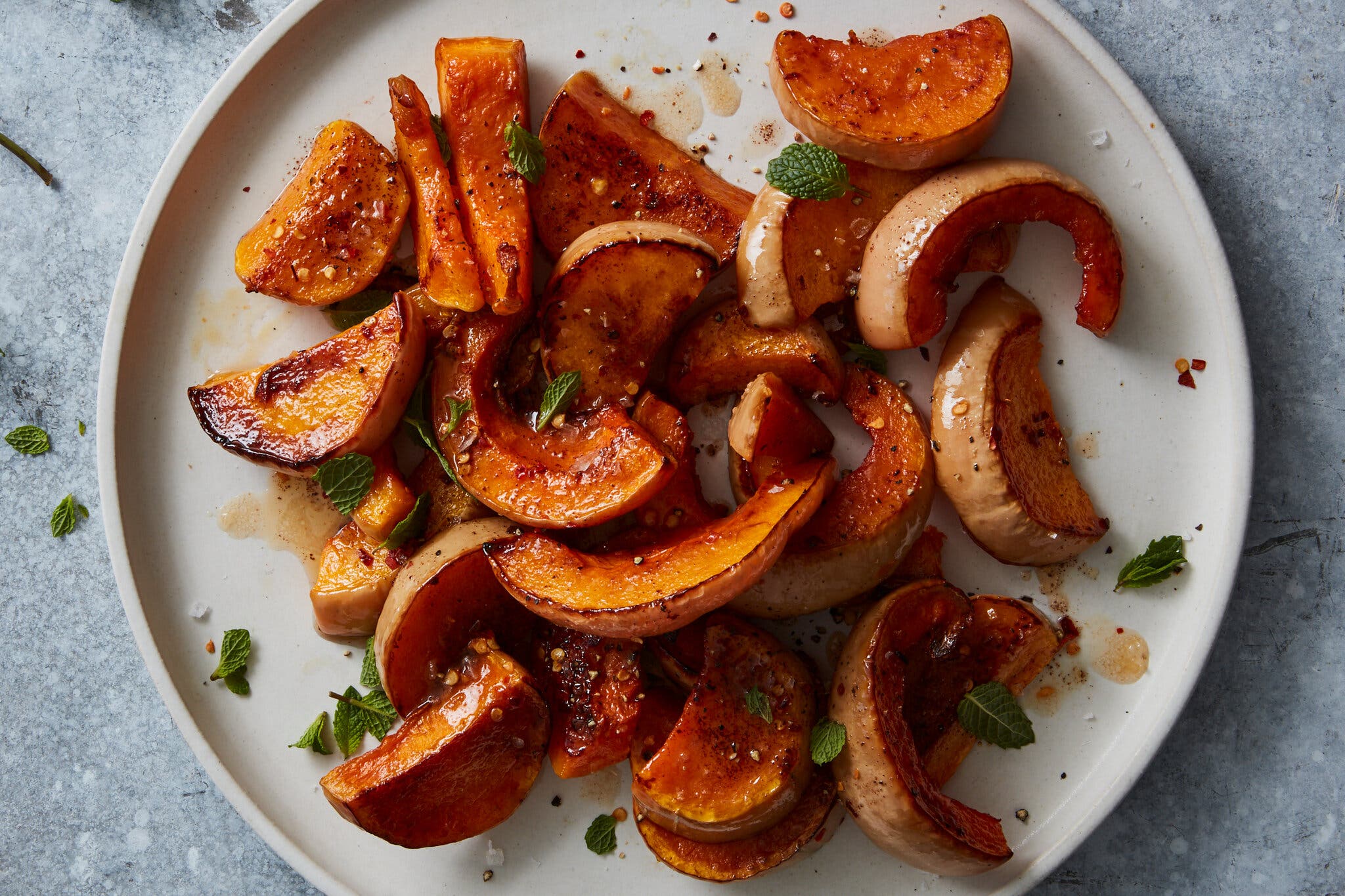 A white plate of roasted butternut squash slices topped with browned butter and herbs sits against a mottled dark gray background.